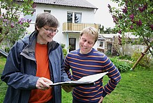 Energieberater mit Unterlagen und Hauseigentümer im Garten, im Hintergrund ein zweigeschossiges Haus