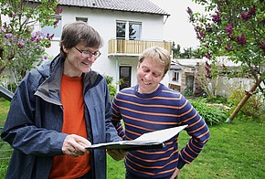 Energieberater mit Unterlagen und Hauseigentümer im Garten, im Hintergrund ein zweigeschossiges Haus